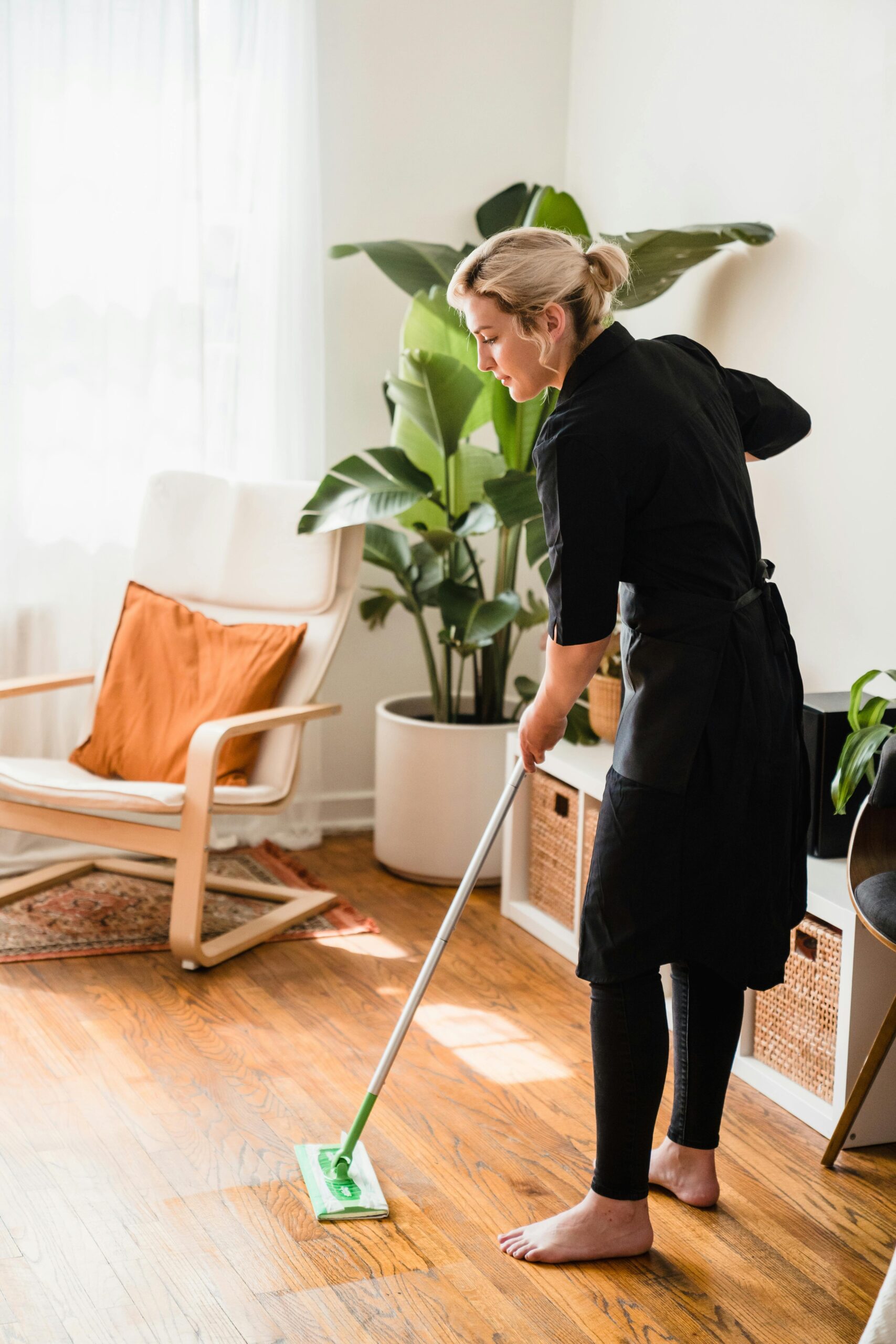 A lady mopping the hard floor with a big green plant in the back.