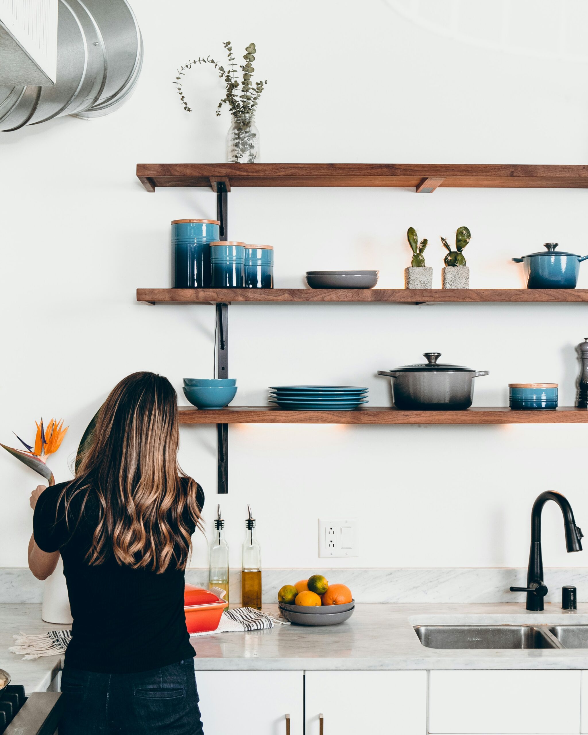 A woman putting flowers in vase on the kitchen shelf.